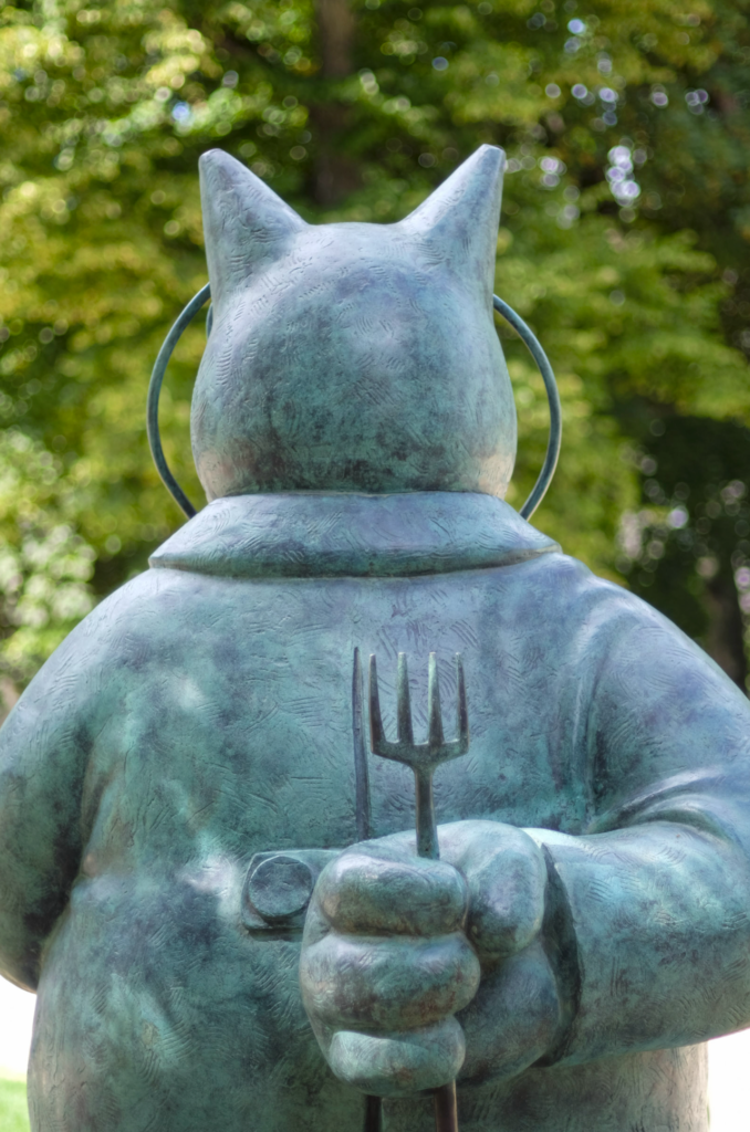 The image features a large statue of a cat holding a fork and knife in its paws. The cat statue is positioned in the middle of the scene, with the fork and knife held prominently in its hands. The statue appears to be made of metal, giving it a unique and artistic appearance. In the background, there are trees visible, adding a natural element to the scene. The combination of the cat statue and the surrounding environment creates an interesting and visually appealing composition.